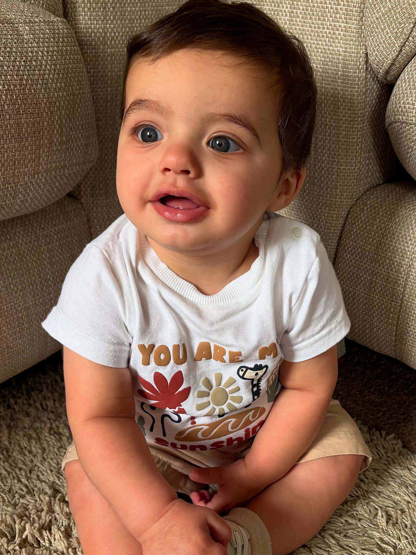Zak joined the competition — help win amazing prizes! toddler, child, baby, sitting, carpet, couch, white_tshirt, shorts, indoor, cute, blue_eyes, curly_hair, expression, hands, legs, smile, plush_furniture, casual_clothing, portrait, young