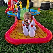 Cain is registered to the contest to win money with this photo: child, toddler, inflatable, water_slide, bowling_pins, outdoor, grass, backyard, play, summer, toy, yellow, red, blue, green, smile, curly_hair, water, fun, sunlight