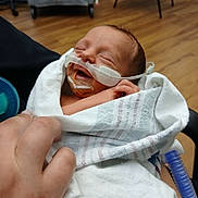 Raymond is registered to the contest to win money with this photo: newborn, baby, infant, blanket, hospital, medical_tube, sleeping, hand, person, healthcare, care, wrapped, face, peaceful, wood_floor, chair, medical_equipment, closeup, indoors, patient