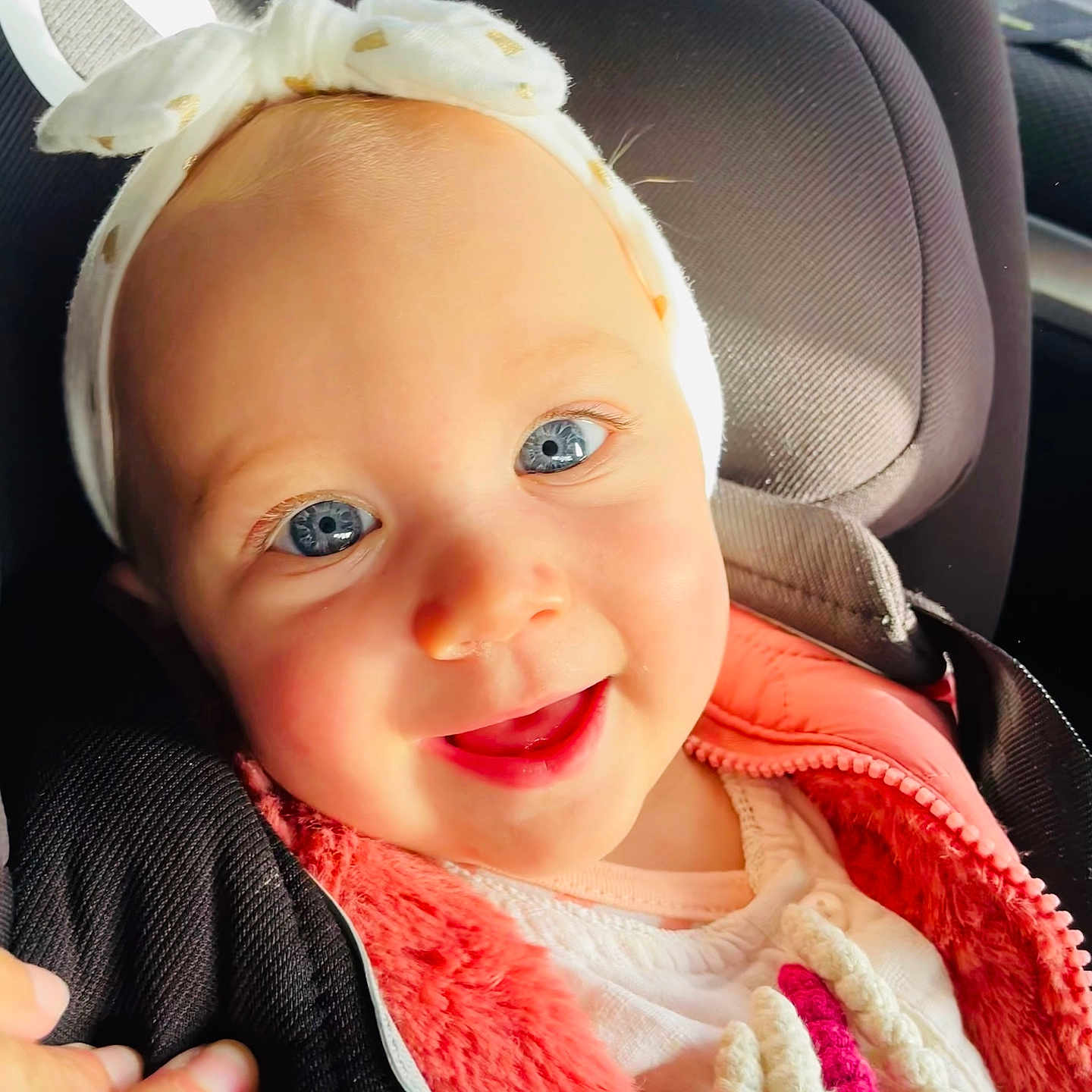 Mïa participe au concours pour gagner de l'argent avec cette photo : baby, blue_eyes, car_seat, child, closeup, cute, face, hand, happy, headband, human, indoors, infant, pink_jacket, portrait, seatbelt, smiling, sunlight, warm_clothing, white_clothing
