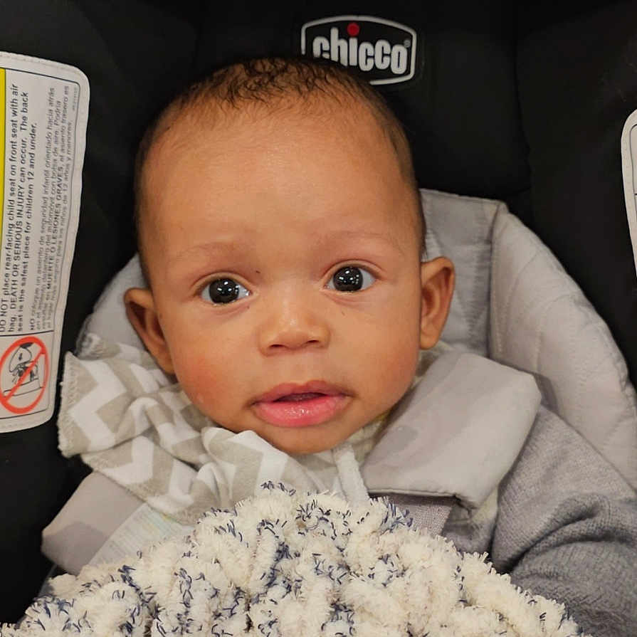 Delonte is registered to the contest to win money with this photo: baby, blanket, bundle, car_seat, child, clothing, cute, expression, eyes, fabric, face, head, indoor, infant, person, portrait, seat, texture, warm, young_child