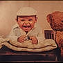 baby, child, smiling, laughing, cap, vest, blanket, bed, teddy_bear, stuffed_animal, wooden_bed, cute, portrait, indoor, happy, cozy, infant, young_child, clothing, soft_lighting