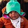 baby, child, hat, turquoise, orange_sweater, bib, dinosaur_print, cute, warm_clothing, outdoor, portrait, face, curious, eyes, headwear, knitwear, infant, person, cozy, winter