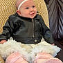 baby, infant, leather_jacket, pink_booties, headband, couch, pillow, smile, blue_eyes, sitting, indoor, clothing, cute, portrait, child, soft_toys, fashion, leather, happy, cozy