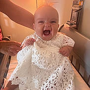 Nova Brielle is registered to the contest to win money with this photo: baby, child, smiling, white_dress, lace, bed, lamp, indoor, person, happy, supporting_hand, infant, cute, portrait, human, clothing, furniture, warm_light, expression, sitting