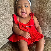 Alani Janae Herrod joined the competition — help win amazing prizes! baby, brown_couch, child, comfortable, couch, cute, flower, girl, happy, headband, indoors, infant, person, portrait, red_dress, sitting, smile, textured_fabric, tongue_out, young_child