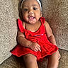 baby, brown_couch, child, comfortable, couch, cute, flower, girl, happy, headband, indoors, infant, person, portrait, red_dress, sitting, smile, textured_fabric, tongue_out, young_child