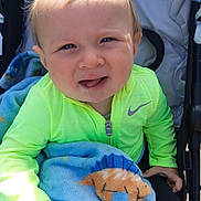 Leo is registered to the contest to win money with this photo: baby, child, stroller, blanket, dinosaur_pattern, blue_blanket, green_jacket, outdoor, cute, infant, person, clothing, hand, face, portrait, young_child, bright_colors, zipper, expression, seated