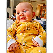 Riverlyn is registered to the contest to win money with this photo: baby, child, infant, smiling, happy, portrait, face, eyes, cheeks, yellow_outfit, floral_pattern, jumpsuit, hands, sitting, indoor, home, bedroom, furniture, close_up, photography