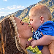 Dallas is registered to the contest to win money with this photo: baby, mother, child, kiss, portrait, outdoor, mountains, landscape, family, parenting, infant, blue_shirt, smiling, affection, sunlight, nature, sky, close_up, 抱抱, travel