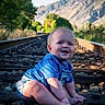 Dallas is registered to the contest to win money with this photo: baby, child, infant, smiling, happy, portrait, sitting, outdoors, railway, railroad_track, train_tracks, mountains, mountain_landscape, greenery, trees, blue_shirt, shorts, sunlight, scenic, nature