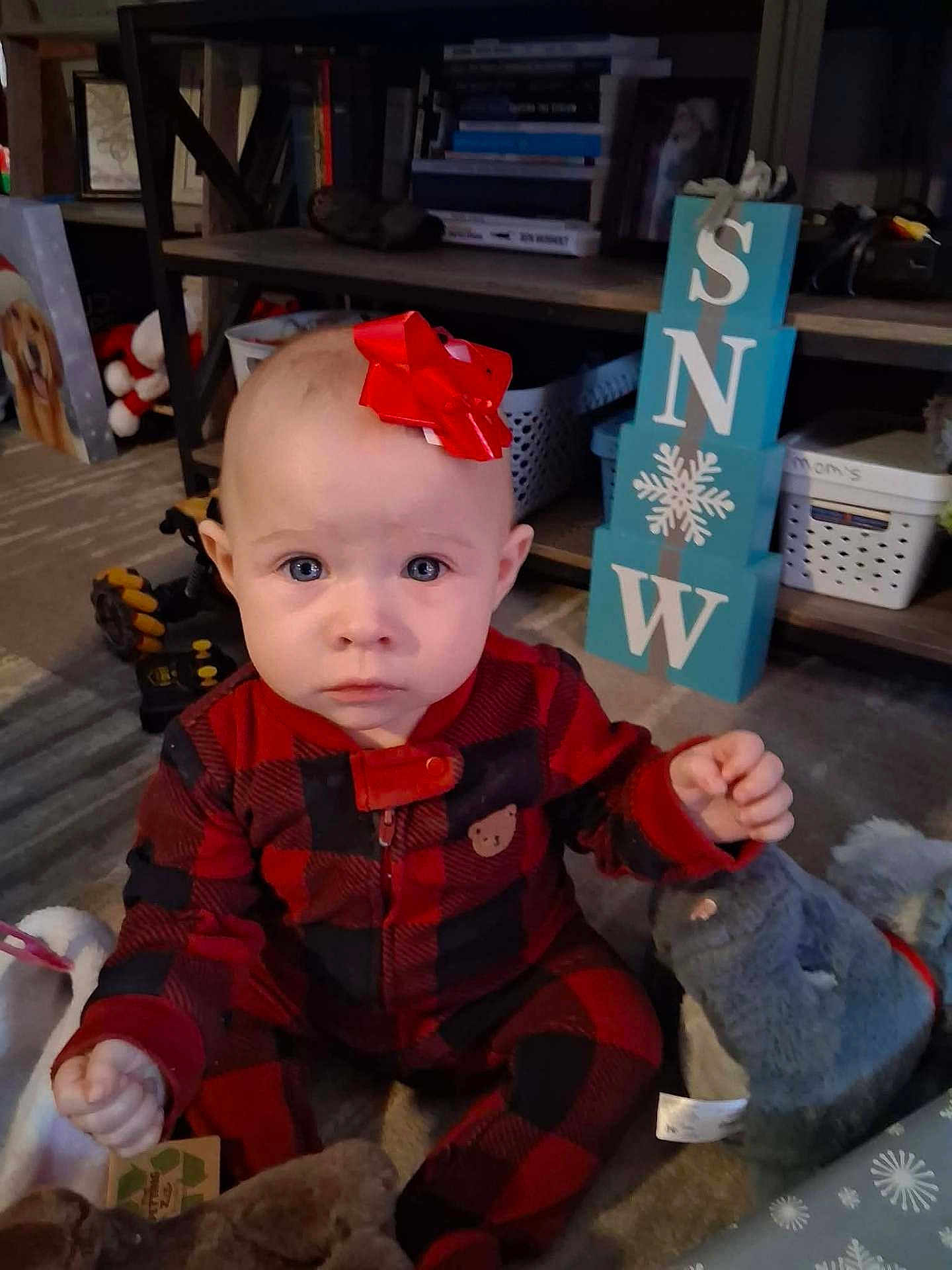 Grace joined the competition — help win amazing prizes! baby, child, indoor, red_bow, plaid_onesie, carpet, toy, stuffed_animal, blue_eyes, holiday_decor, shelf, books, basket, floor, person, cute, infant, winter, home, curious