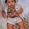 baby, child, infant, portrait, sitting, lying_down, fashion, designer_clothing, hat, bucket_hat, sunglasses, necklace, gold_chain, sandals, bed, blanket, indoors, smiling, luxury, gucci