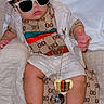 baby, child, infant, portrait, sitting, sunglasses, bucket_hat, hat, fashion, outfit, white_clothing, sandals, pacifier, blanket, bed, indoor, smiling, face, jewelry, accessories