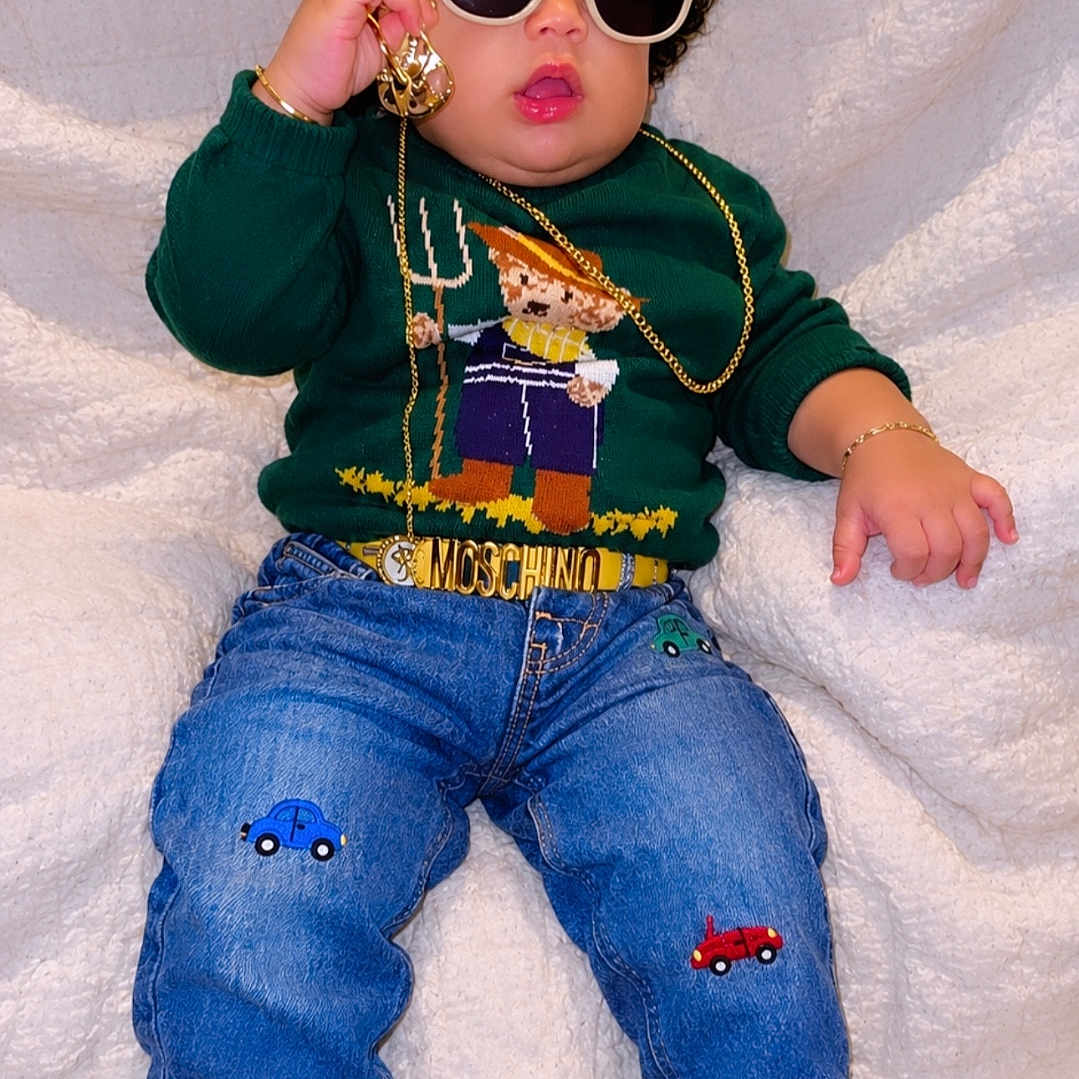 Kayden is registered to the contest to win money with this photo: child, baby, toddler, portrait, sitting, fashion, sunglasses, jewelry, sweater, jeans, sneakers, green, black, gold, curly_hair, fluffy_blanket, indoor, clothing, smiling, stylish