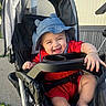 baby, black_stroller, blue_hat, bracelet, child, cute, hand, happy, hat, infant, joyful, legs, outdoor, person, playful, red_clothing, seatbelt, smiling, stroller, sunlight