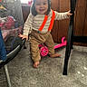 toddler, child, standing, smiling, portrait, indoors, suspenders, striped_shirt, brown_pants, barefoot, pink_scooter, furniture, desk, carpet, window, playroom, trampoline, happy, young_child, home