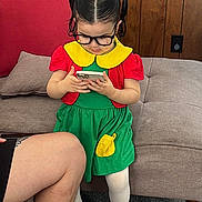 Alaia is registered to the contest to win money with this photo: child, girl, toddler, glasses, pigtails, smartphone, indoors, couch, wood_paneling, colorful_clothing, dress, red_shirt, green_skirt, yellow_collar, white_tights, black_shoes, carpet, portrait, sitting_area, adult_leg