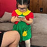 child, girl, toddler, glasses, pigtails, smartphone, indoors, couch, wood_paneling, colorful_clothing, dress, red_shirt, green_skirt, yellow_collar, white_tights, black_shoes, carpet, portrait, sitting_area, adult_leg