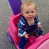 blue_eyes, casual_clothing, child, cute, daylight, denim_overalls, face, happy, long_sleeves, outdoor, pavement, person, pink, playing, purple, small_vehicle, steering_wheel, toddler, toy_car, young_child