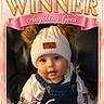 baby, child, hat, knit_hat, pom_poms, denim_overalls, cute, portrait, face, blue_eyes, pink_border, crown, text_overlay, indoor, seated, adorable, clothing, person, smiling, warm_lighting