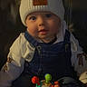 baby, child, clothing, colorful_toy, cute, denim_overalls, face, hat, headwear, indoor, infant, leather_chair, playful, pom_poms, portrait, seated, smiling, toy, warm_lighting, white_shirt