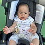 baby, stroller, infant, child, clothing, pajamas, hands, feet, face, expression, seat, safety_belt, toy, pacifier_clip, outdoor, daylight, green_background, comfort, cute, curious