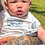 baby, child, tattoo, arm, hand, shirt, white_shirt, shark_print, outdoor, sunlight, grass, person, portrait, skin, expression, holding, casual_clothing, daytime, nature, closeup