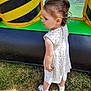 toddler, child, white_dress, grass, outdoor, inflatable, playground, curious, side_view, summer, shoes, patterned_clothing, yellow, black, green, hair_tied, young_child, standing, sunlight, play