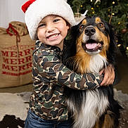 Sadie joined the competition — help win amazing prizes! child, dog, smiling, santa_hat, christmas_tree, holiday, festive, camouflage_clothing, hug, indoor, decorations, lights, joy, friendship, fur, kneeling, happy, canine, celebration, portrait