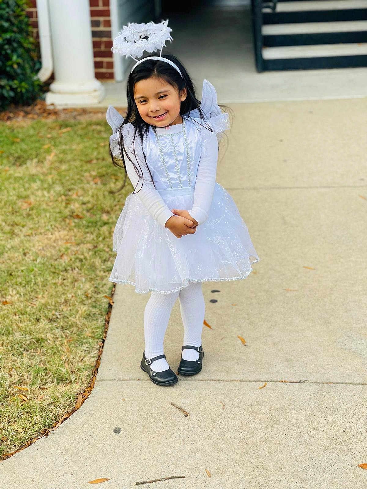 Victoria Huerta is registered to the contest to win money with this photo: child, girl, angel_costume, white_dress, halo, tights, black_shoes, sidewalk, grass, smiling, long_hair, pose, outdoor, daylight, costume, cute, portrait, holiday, happy, fashion
