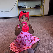 Naomi is registered to the contest to win money with this photo: toddler, child, girl, pink_dress, red_bow, bottle, carpet, indoor, television, furniture, sitting, drinking, headband, cute, young_child, floor, toy, entertainment, electronics, home