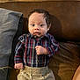 baby, beige_pants, brown_couch, casual_clothing, child, curious, cute, face, gray_pillow, hands, home, indoor, infant, person, plaid_shirt, portrait, relaxed, seated, wide_eyes, young_child