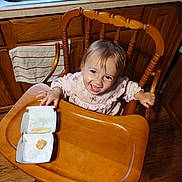 Annabelle is registered to the contest to win money with this photo: child, toddler, baby, smiling, happy, portrait, high_chair, kitchen, wooden_furniture, snack, food, takeout_box, tray, dress, indoor, sink, cabinets, floor, seated, face