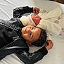 child, baby, sibling, bed, white_bed_sheet, black_jacket, pajamas, hat, smile, lying_down, cozy, cute, indoors, infant, warm_clothing, person, sleeping_baby, soft_toys, family, toddler