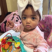 Elena is registered to the contest to win money with this photo: baby, child, high_chair, toy, headband, bow, pink_clothing, wooden_toy, beads, orange, indoor, blanket, colorful, infant, cute, playtime, person, portrait, curious, seated