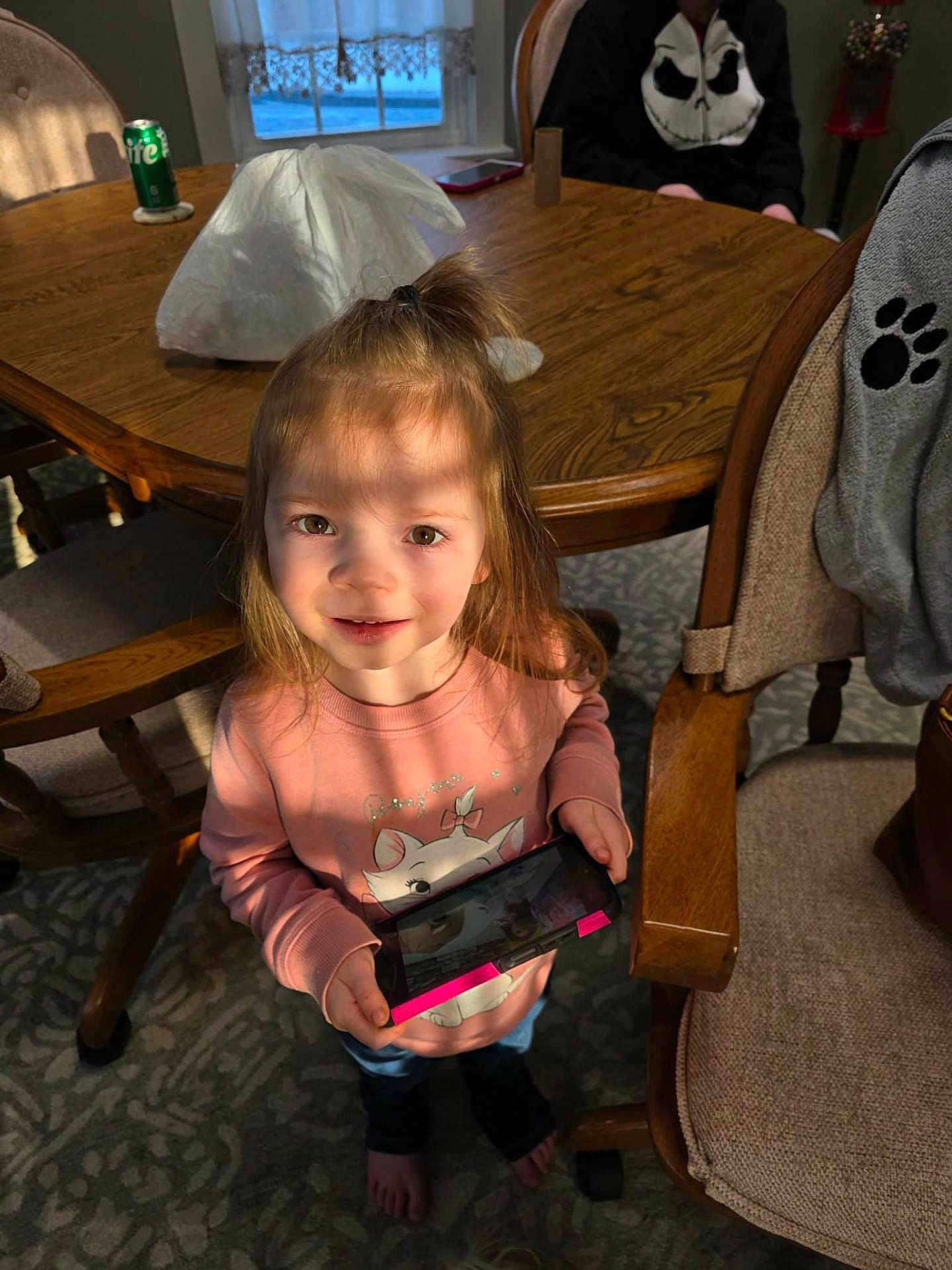 Aaliyah joined the competition — help win amazing prizes! child, girl, indoors, table, chair, wooden_furniture, carpet, handheld_device, pink_sweater, barefoot, sunlight, shadow, smiling, person, casual_clothing, living_room, curtain, window, beverage_can, playing