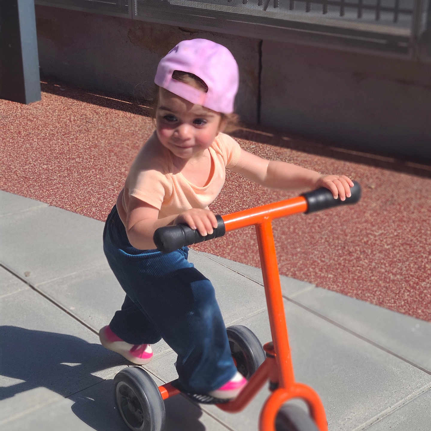 Lia Yasemin a rejoint le concours — aidez-le/la à gagner de superbes lots ! active, blue_pants, casual_clothing, child, daytime, fun, orange_scooter, outdoor, pavement, person, pink_cap, play, playground, scooter, shadow, shoes, smiling, sunlight, toddler, young_child
