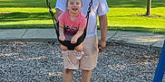 Saylor is registered to the contest to win money with this photo: adult, casual_clothing, child, daylight, family, fun, grass, happy, nature, outdoor, park, person, playground, recreation, sandals, shorts, smiling, swing, toddler, tree