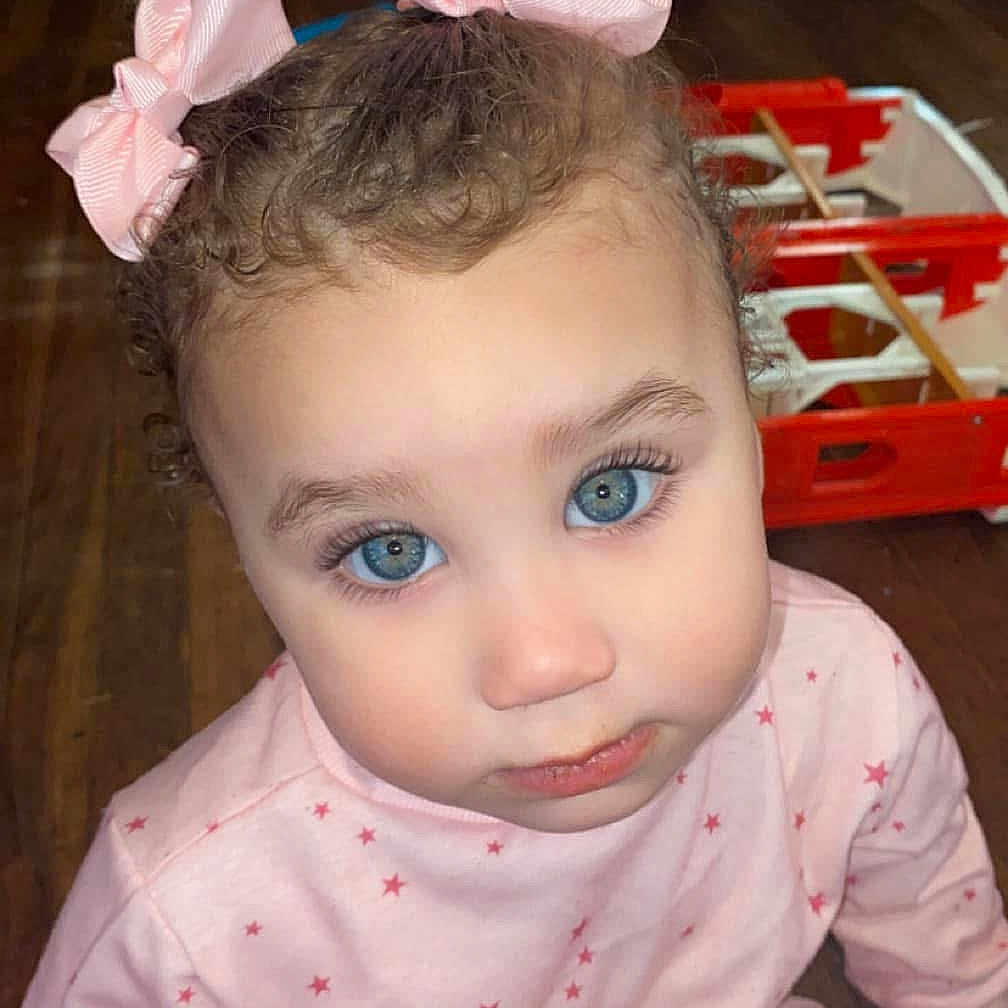 Saylor is registered to the contest to win money with this photo: baby, blue_eyes, child, closeup, curious, curly_hair, cute, face, home, indoor, looking_up, pajamas, person, pink_bows, sitting, stars_pattern, toddler, toy, wooden_floor, young_child