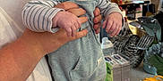 Vinnie is registered to the contest to win money with this photo: adult_hand, baby, background, black_shoes, child, countertop, cute, decor, holding, household_items, indoor, infant, kitchen, overalls, pacifier, person, striped_clothing, table, toy, window