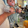 Vinnie is registered to the contest to win money with this photo: baby, pacifier, child, striped_clothing, overalls, black_shoes, adult_hand, kitchen, countertop, household_items, window, indoor, person, cute, infant, holding, table, toy, background, decor