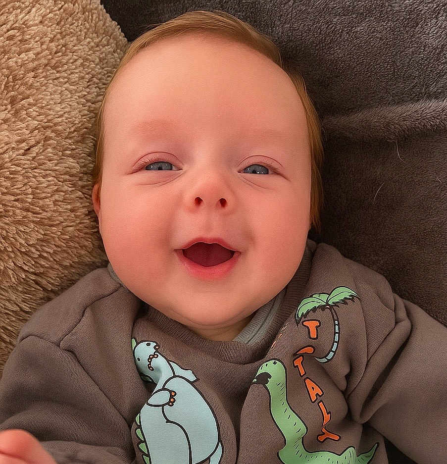 Vinnie is registered to the contest to win money with this photo: baby, smiling, face, infant, clothing, sweatshirt, dinosaur, blanket, cozy, cute, child, person, happy, head, skin, closeup, indoors, young, apparel, portrait
