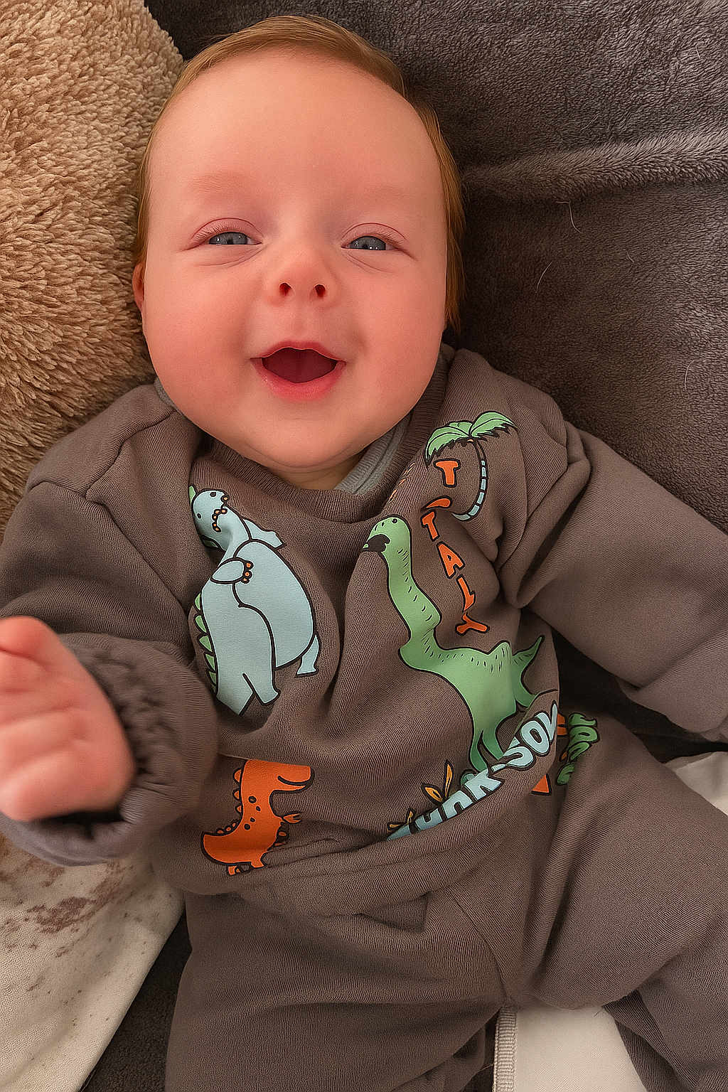 Vinnie is registered to the contest to win money with this photo: baby, smiling, infant, dinosaur_clothing, cozy, cute, happy, child, blanket, plush, indoors, portrait, face, clothing, sleeves, hand, head, lying_down, soft_texture, young