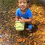 toddler, child, water, stream, rocks, bucket, blue_shirt, wet_hair, smiling, outdoor, nature, sandals, playful, sunlight, happy, sitting, water_reflection, summer, daylight, cute