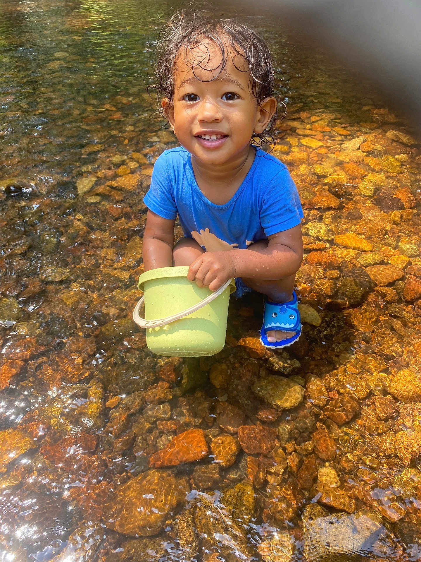 Keydji participe au concours pour gagner de l'argent avec cette photo : toddler, child, water, stream, rocks, bucket, blue_shirt, wet_hair, smiling, outdoor, nature, sandals, playful, sunlight, happy, sitting, water_reflection, summer, daylight, cute