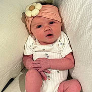 Eliana is registered to the contest to win money with this photo: baby, infant, headband, flower, couch, cushion, white_clothing, sitting, hands, legs, skin, face, expression, indoors, soft, comfortable, child, portrait, cute, small