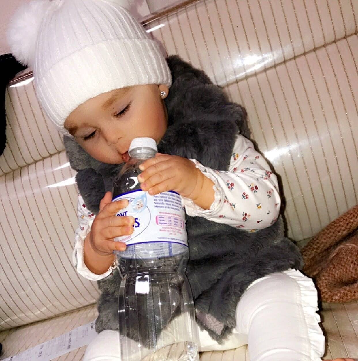 Chelsea participe au concours pour gagner de l'argent avec cette photo : beanie, black_hair, bottle, cap, car_seat, drink, drinking, drinkware, fur, glass_bottle, hand, headwear, knit_cap, long_hair, nail, person, plastic_bottle, room, sitting, textile