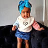 black_sofa, blue_bow, child, colorful_clothing, curly_hair, cute, denim_skirt, fashion, headband, heart_design, home, indoor, person, playful, standing, sweater, toddler, wall, white_shoes, young_child