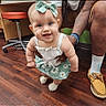 baby, child, infant, bow, green_bow, dress, floral_dress, smile, tattoo, adult_hand, wooden_floor, indoor, footwear, loafers, white_socks, cute, happy, supporting_hand, person, flooring